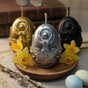 Peut inclure: Trois bougies en forme d'œuf, dorées, argentées et noires, avec des motifs en relief complexes. Des jonquilles jaunes et des œufs décoratifs entourent les bougies, disposées sur une assiette en bois et un tissu à motifs.