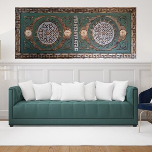 May include: A teal green sofa with white cushions sits in front of a wall with a wooden carved wall art piece with a green and brown background. The wall art features a geometric design with a silver and white colour scheme.