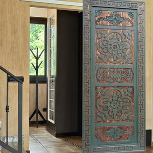 May include: A wooden door with intricate carvings and a green paint finish. The door has a Greek key design border and features a repeating floral pattern.