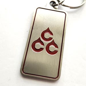 May include: Silver rectangular keychain with a red border and a red emblem of three connected C shapes. The keychain has a silver key ring attached at the top.