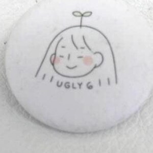 May include: A white button with a hand-drawn illustration of a smiling face with a sprout on top. The face has pink cheeks and the text "UGLY 611" is written below the face in black.