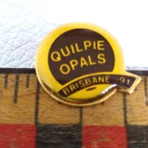Może przedstawiać: Żółto-brązowa emaliowana przypinka z napisem "QUILPIE OPALS BRISBANE 91". Przypinka jest okrągła z przedłużeniem w kształcie banera. Przypinka jest umieszczona na drewnianej linijce.