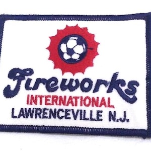 May include: A rectangular patch with a white background, a navy blue border, and navy blue text that reads "Fireworks International Lawrenceville N.J." A red burst design surrounds a soccer ball graphic.