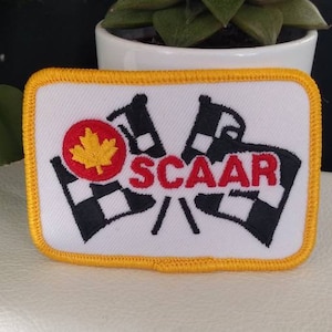 Puede incluir: Un parche bordado blanco con una hoja de arce roja, dos banderas a cuadros blanco y negro y el texto "SCAA" en rojo. El parche tiene un borde amarillo.