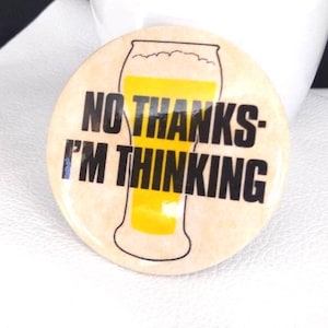 May include: A round button with the text "NO THANKS - I'M THINKING" in black, with a yellow beer glass graphic. The button has a light tan background and is set against a white surface with a white ceramic pot and green plant in the background.