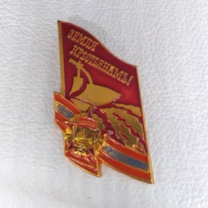 May include: A red and gold enamel pin with a red flag that says "Земля крестьянам!" in gold lettering. The flag has a gold sickle and hammer on it. The pin also has a gold star and a gold ribbon.