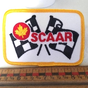 May include: Rectangular patch with a white background, featuring two checkered flags and the word "SCAAR" in red. A red circle with a yellow maple leaf is on the left. The patch has a yellow border.