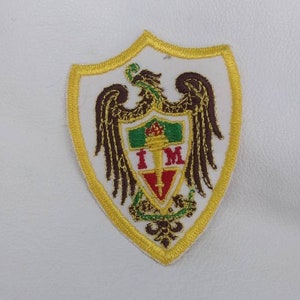 Può includere: Toppa ricamata con un disegno a scudo. La toppa ha uno sfondo bianco e un bordo dorato. Lo scudo contiene un'aquila marrone, dettagli verdi e rossi e le lettere "I M".