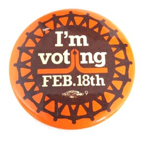 Puede incluir: Botón redondo naranja con el texto "I'm voting FEB.18th" en blanco y marrón. El botón tiene un centro marrón con un borde naranja decorativo. Un botón de campaña política vintage.