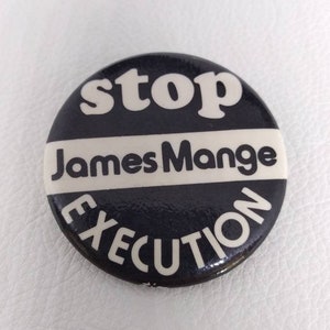 May include: A round, black and white button with the words "stop James Mange EXECUTION" in white lettering. The button has a glossy finish and is set against a white background.