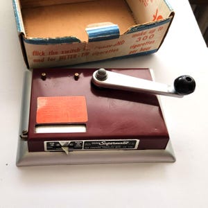 May include: A vintage Supermatic cigarette rolling machine with a maroon body, silver base, and a black handle. The machine is accompanied by its original cardboard box. The box has text that reads "flick the switch...and for FILTER-TIP cigarettes."