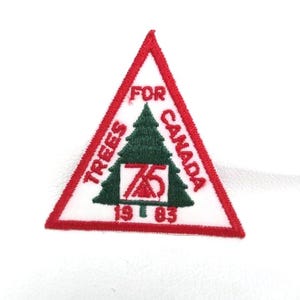May include: A triangular patch with a red border and white background. The patch features the text "TREES FOR CANADA" in red, surrounding a green Christmas tree and the number "75" in red. The year "1983" is at the bottom.