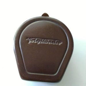 Puede incluir: Tapa de objetivo de cámara de cuero marrón con el nombre de la marca "Voigtländer" en relieve. La tapa tiene forma rectangular redondeada con una pequeña presilla de cuero. Fondo blanco.