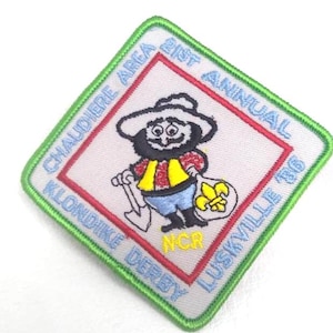 May include: A square patch with a green border featuring a cartoon prospector with a pickaxe and a bag of gold nuggets. The patch reads "21st Annual Klondike Derby Luskville '86" and "Chaudiere Area" in blue text.