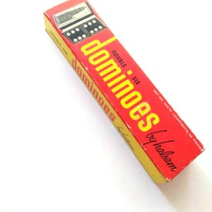 Op de afbeelding: Een vintage dominostenenset in een rood en gele doos. De doos toont het woord "dominoes" in grote gele letters, met "DOUBLE SIX" aan de zijkant. De doos heeft ook een illustratie van dominostenen.