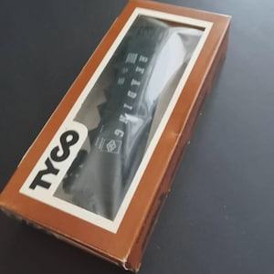 May include: A brown rectangular box with a clear window displaying a black object. The box has the word "TYCO" in white and the word "READING" printed on the object. The box is on a dark surface.