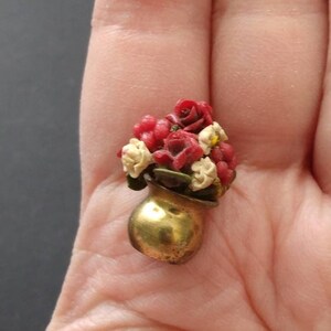 May include: Miniature gold-colored vase filled with red and cream-colored roses. The small bouquet includes green leaves and yellow accents. The vase has a rounded shape and a slightly textured surface. The overall theme is floral and decorative.