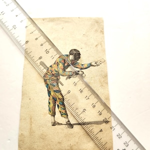 May include: An antique print of a figure in a colorful harlequin costume, holding a small object. The figure is depicted in a dynamic pose, with a sword at their side. The print is on aged paper, with a ruler partially obscuring the image.