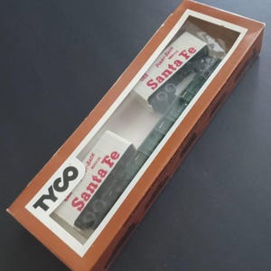 May include: A vintage Tyco Santa Fe model train set in its original box. The box is brown with a clear window, displaying the green train cars and the Santa Fe logo. The box is rectangular and angled.