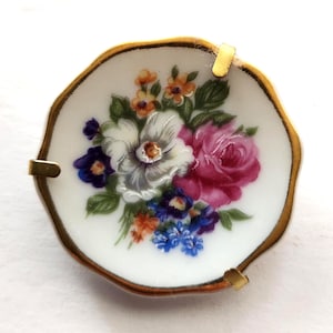 May include: A round, white porcelain brooch with a gold-colored edge. The brooch features a floral design with a large pink rose, white flower, and blue and orange accent flowers. Two gold-colored metal clasps are attached.