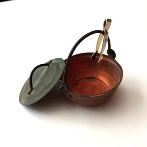 May include: A miniature copper-coloured cauldron with a dark handle and lid. The lid is slightly ajar, revealing the interior. A small, gold-coloured pair of tongs hangs from the handle. The cauldron is likely a decorative item or a miniature cooking pot.