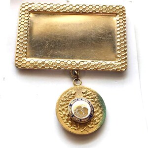 Puede incluir: Un broche vintage dorado con una parte superior rectangular y un colgante circular. La parte superior rectangular tiene un borde texturizado en forma de panal. El colgante presenta texto y un diseño dentro de un borde azul y blanco, con un fondo verde y dorado.
