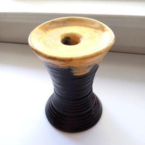 May include: A handmade ceramic candle holder with a unique hourglass shape. The top is a light tan color, transitioning to a dark brown base with a textured, ridged design. The candle holder has a hole in the center.