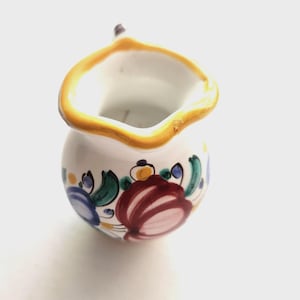 Könnte beinhalten: Ein kleiner weißer Keramikkrug mit einem handgemalten Blumenmuster aus roten, blauen und grünen Blütenblättern. Der Rand des Krugs ist mit einer dicken gelben Umrandung akzentuiert und der Körper hat eine abgerundete Form mit einem kleinen Ausguss.