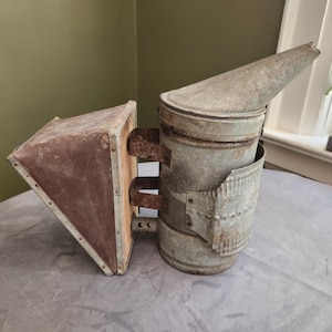 Puede incluir: Un ahumador de abejas de metal vintage que cuenta con un recipiente cilíndrico con boquilla cónica y un fuelle. El dispositivo tiene un acabado desgastado y oxidado con óxido visible en los componentes metálicos y material marrón texturizado en el fuelle.