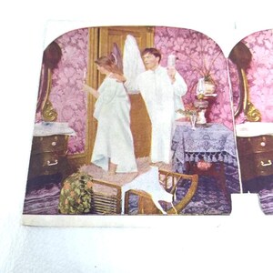 May include: A vintage stereoscopic card depicting a domestic scene. Two figures in white robes stand in a room with pink floral wallpaper. A table with a lace cloth and a dresser are also visible. The card is in a curved shape.