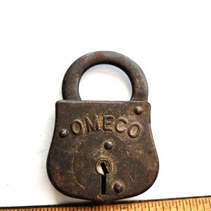 Puede incluir: Un candado de metal vintage y desgastado con un asa curva. La palabra "OMECO" está grabada en la parte delantera. El candado es de color marrón oscuro con signos de edad y desgaste. Una regla de madera está en primer plano.