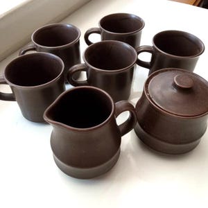 May include: A set of brown ceramic mugs, a creamer, and a sugar bowl with a lid. The mugs have a simple, rounded design with handles. The creamer has a spout and handle. The sugar bowl is round with a small knob on the lid.