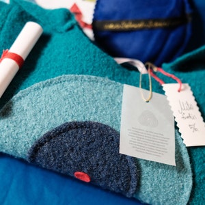 May include: A teal blue sweater with a large, dark blue circle design. The sweater has a white tag with text about the material and a smaller white tag with the text "Mille Grazie 56."