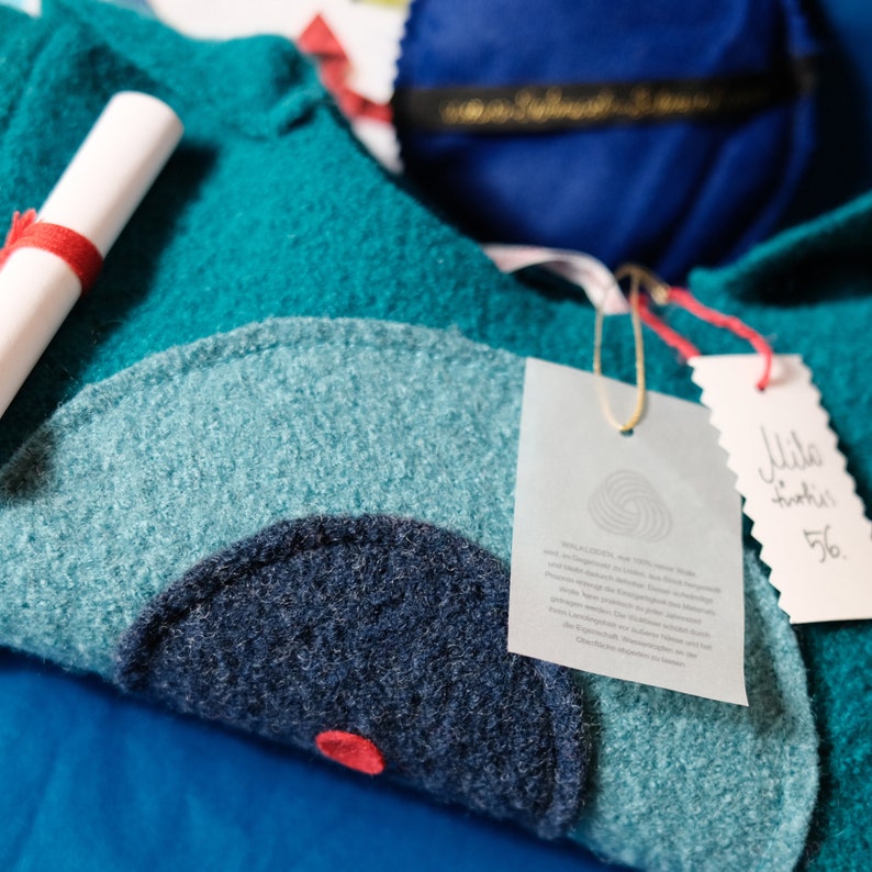 May include: A teal blue sweater with a large, dark blue circle design. The sweater has a white tag with text about the material and a smaller white tag with the text "Mille Grazie 56."