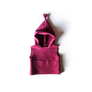 Könnte beinhalten: Eine ärmellose Kapuzenweste in Magenta mit zwei abgerundeten Taschen. Die Kapuze ist spitz und hat ein geknotetes Detail oben. Die Weste ist aus einem weichen, filzartigen Material gefertigt.
