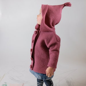 以下が含まれることがあります： フードに小さなタッセルが付いたピンクのフード付きジャケットを着た幼児。ジャケットはボタン留めになっており、柔らかくふわふわした素材で作られています。