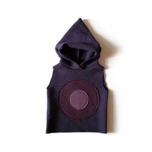 May include: A sleeveless, hooded vest in a deep purple colour. The vest has a large, circular design on the front, with a darker purple circle at the centre. The hood is pointed, and the garment is made of a soft, textured material.