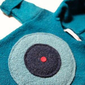 May include: A teal blue hooded sweater with a large, circular, navy blue and light blue eye design. The eye has a red dot in the centre.
