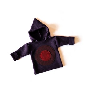 May include: A dark purple hooded sweatshirt with a circular design on the front. The design features a dark red centre circle surrounded by a maroon and dark purple circle. The sweatshirt has long sleeves and a pointed hood.