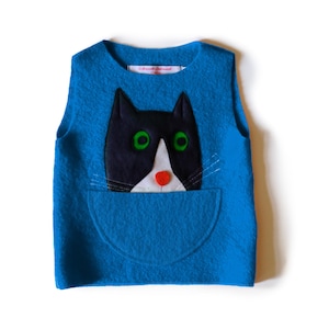 May include: A blue felt vest with a cat design. The cat's face is black and white with green eyes and a red nose, emerging from a pocket. The vest has a round neck and a small tag that reads "schneid schick".