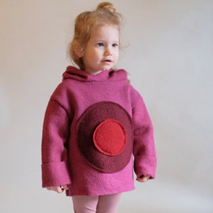 May include: A pink hooded sweatshirt with a large, multi-coloured circle design on the front. The circle is made of felt and has three layers of different shades of red.