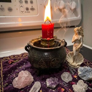 May include: A red candle with a bright flame sits inside a dark, textured metal cauldron. Beside it stands a small, cream-colored figurine of a person emerging from a shell. Several raw crystals, including rose quartz and amethyst, rest on a purple cloth.