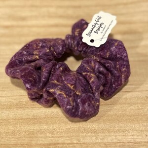 May include: A purple scrunchie with gold flecks. The scrunchie is made of a soft fabric and is gathered to create a voluminous look. A tag with the text "Scrunchy Girl Designs" is attached.