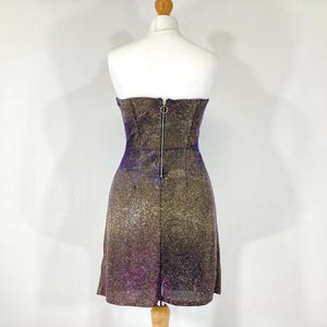 May include: A strapless mini dress on a mannequin. The dress features a shimmering, glittery fabric with a gradient effect, transitioning from purple to gold. A zipper runs down the back. The dress is displayed on a white background.