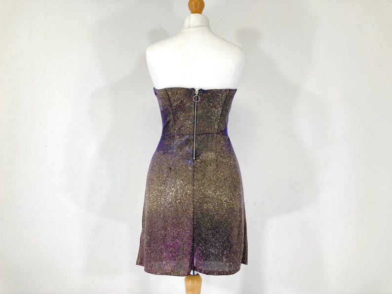 May include: A strapless mini dress on a mannequin. The dress features a shimmering, glittery fabric with a gradient effect, transitioning from purple to gold. A zipper runs down the back. The dress is displayed on a white background.