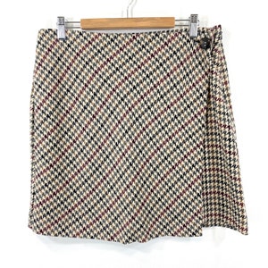 May include: A beige, black, brown, and red houndstooth patterned skirt with a single button closure on the side.