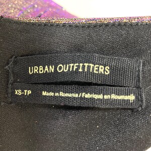 May include: Close-up of a black garment tag with the text "URBAN OUTFITTERS" in white. Below the brand name is "XS-TP" and "Made in Romania / Fabriqu&eacute; en Roumanie". A silver zipper is visible on the left.