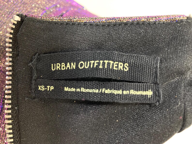 May include: Close-up of a black garment tag with the text "URBAN OUTFITTERS" in white. Below the brand name is "XS-TP" and "Made in Romania / Fabriqu&eacute; en Roumanie". A silver zipper is visible on the left.