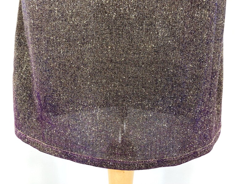 May include: Close-up of a dark purple dress with a shimmering, textured fabric. The dress has a rounded hemline and a subtle sheen, suggesting a formal or evening wear garment. The fabric appears to be a knit or woven material.