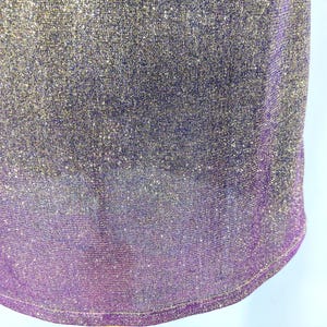 May include: Close-up of a shimmering fabric with a gradient effect. The top portion is a blend of gold and silver, transitioning to a deep purple at the bottom. The fabric has a textured, glittery appearance.